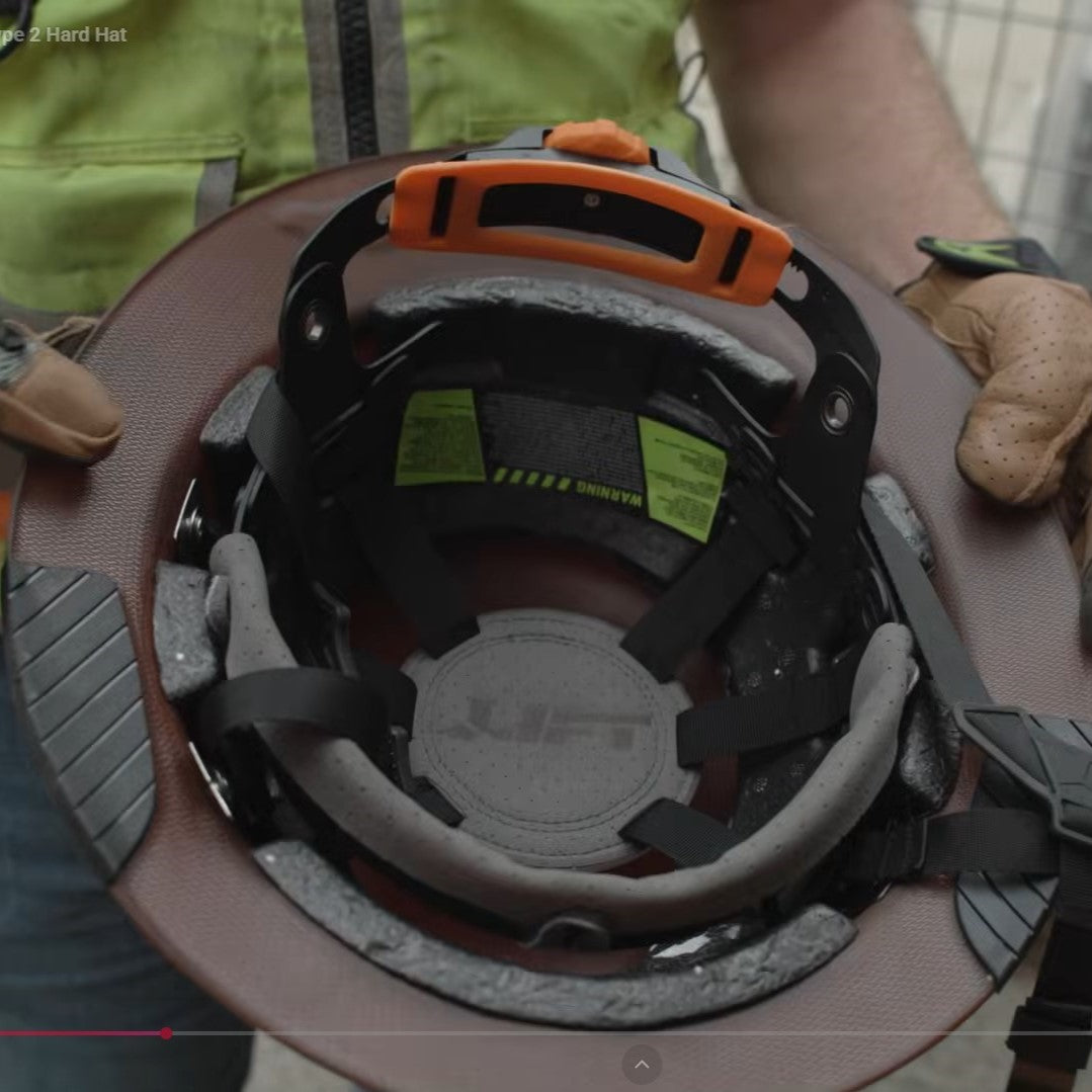Close-up of the inside of a Lift Safety DAX T2 showing the suspension, straps, and a black-and-orange top handle. A person wearing a high-visibility vest and gloves is holding the hat.